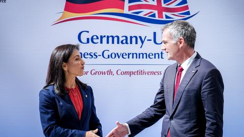 Reiche begrüßt Kyle mit Handschlag. Beide schauen sich an. Im Hintergrund sind die Flaggen beider Länder zu sehen. 