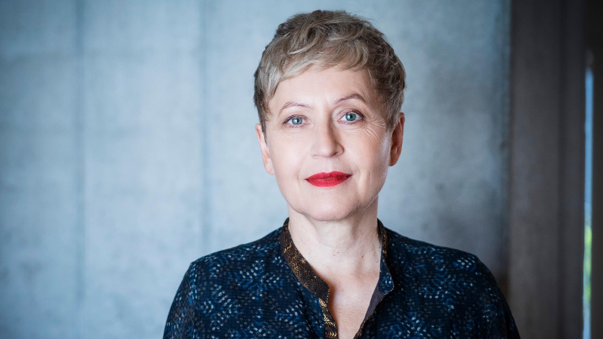 Ulrike Draesner schaut in die Kamera. Sie trägt ein dunkelblaue, gemusterte Bluse. Ihre Haare trägt sie kur, ihre Lippen hat sie rot geschminkt, sie hat blaue Augen.