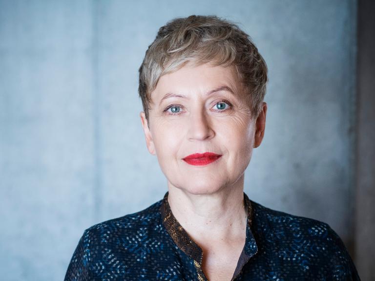 Ulrike Draesner schaut in die Kamera. Sie trägt ein dunkelblaue, gemusterte Bluse. Ihre Haare trägt sie kur, ihre Lippen hat sie rot geschminkt, sie hat blaue Augen.