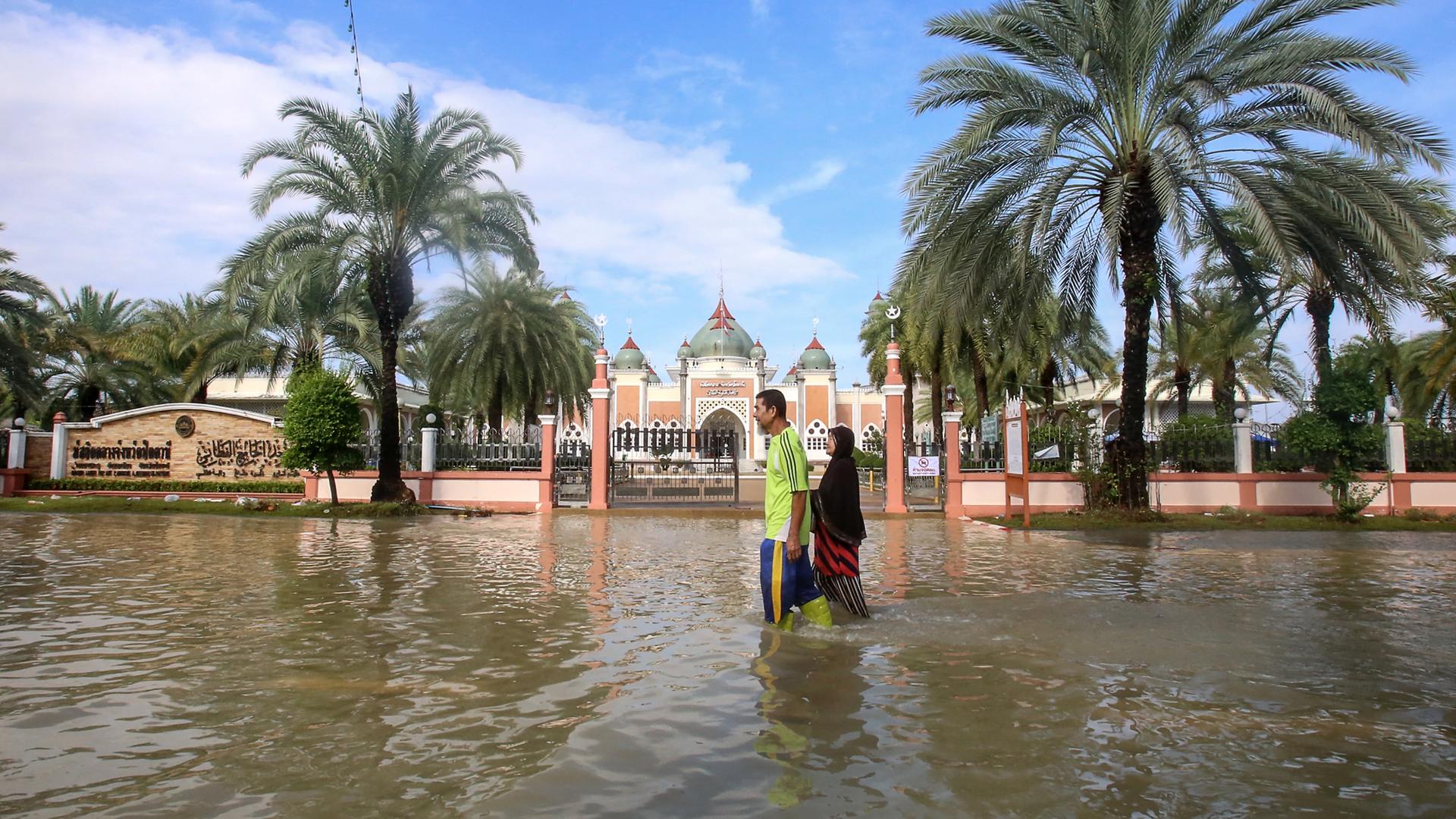 Zwei Personen waten durch kniehohes Wasser über eine Straße außerhalb von Pattani in Thailand.