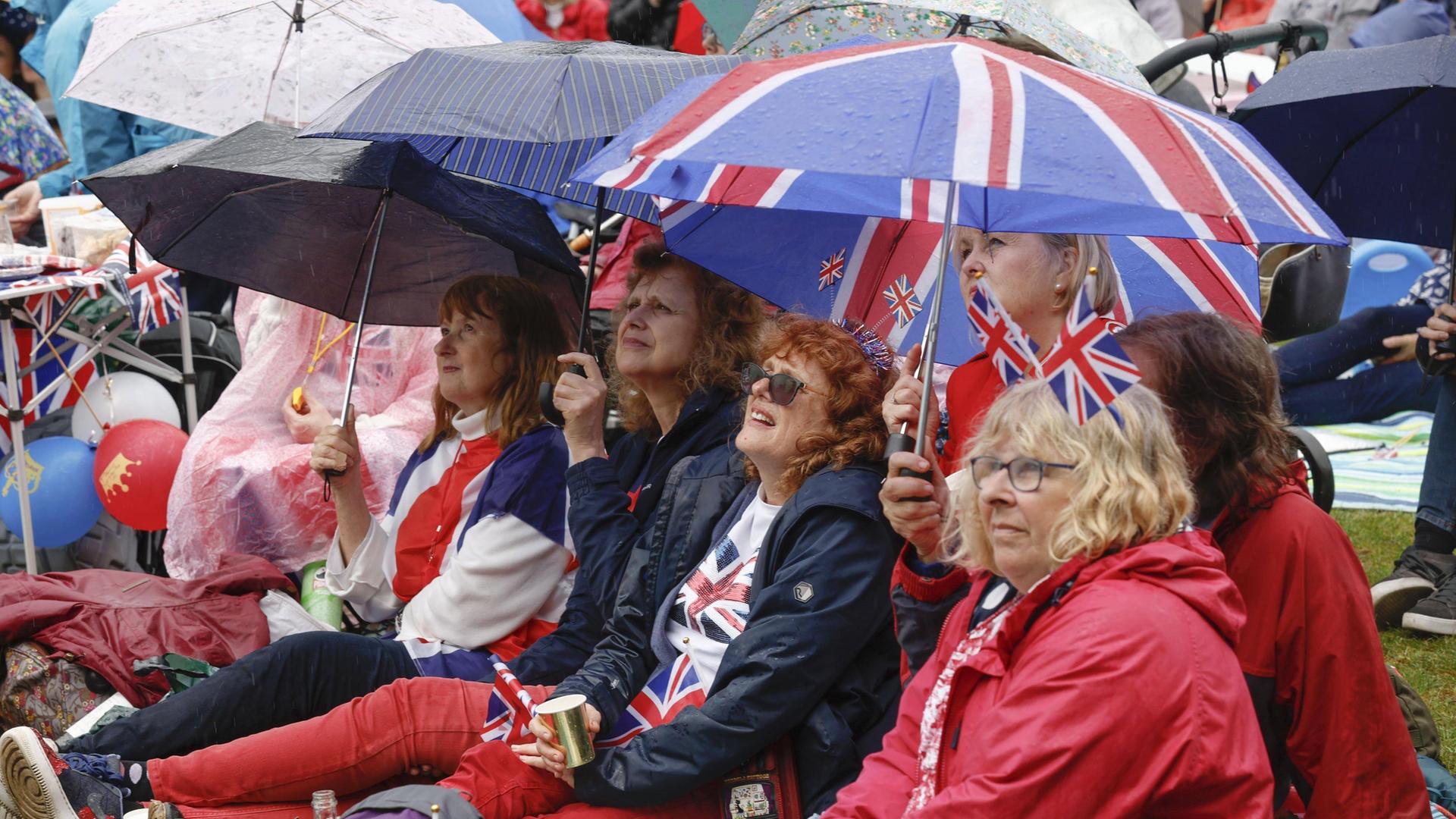 Mehrere Frauen sitzen unter Regenschirmen in den Farben der britischen Flagge auf dem Boden und schauen gemeinsam die Krönung von König Charles III. in einer Videoübertragung.