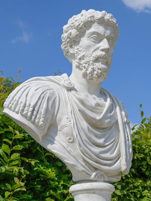 Eine Büste des römischen Kaisers Mark Aurel vor Gebüsch und blauem Himmel.