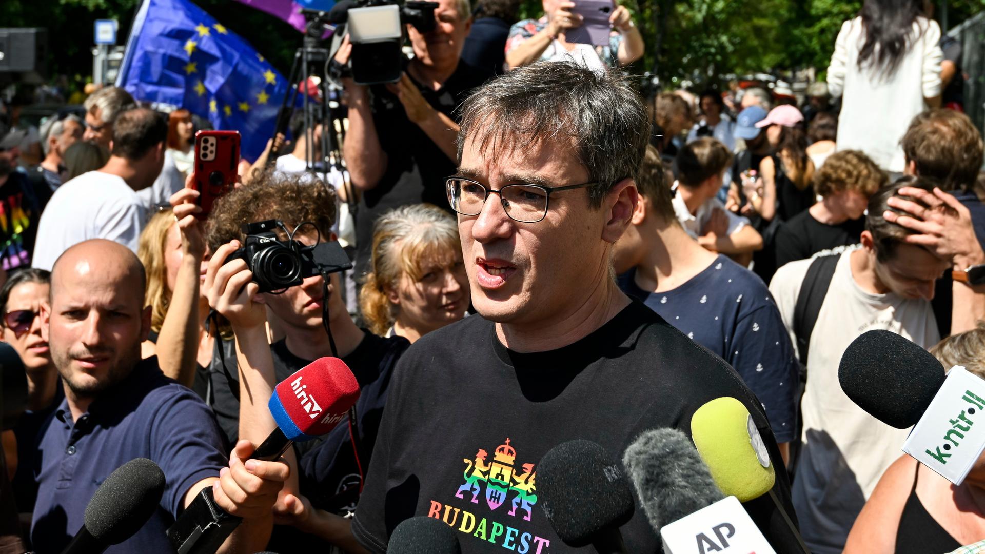 Karacsony steht zwischen Reportern und spricht. Er trägt ein schwarzes Shirrt mit Schrift in den Regenbogenfarben. Dahinter viele Teilnehmer des Marsches.