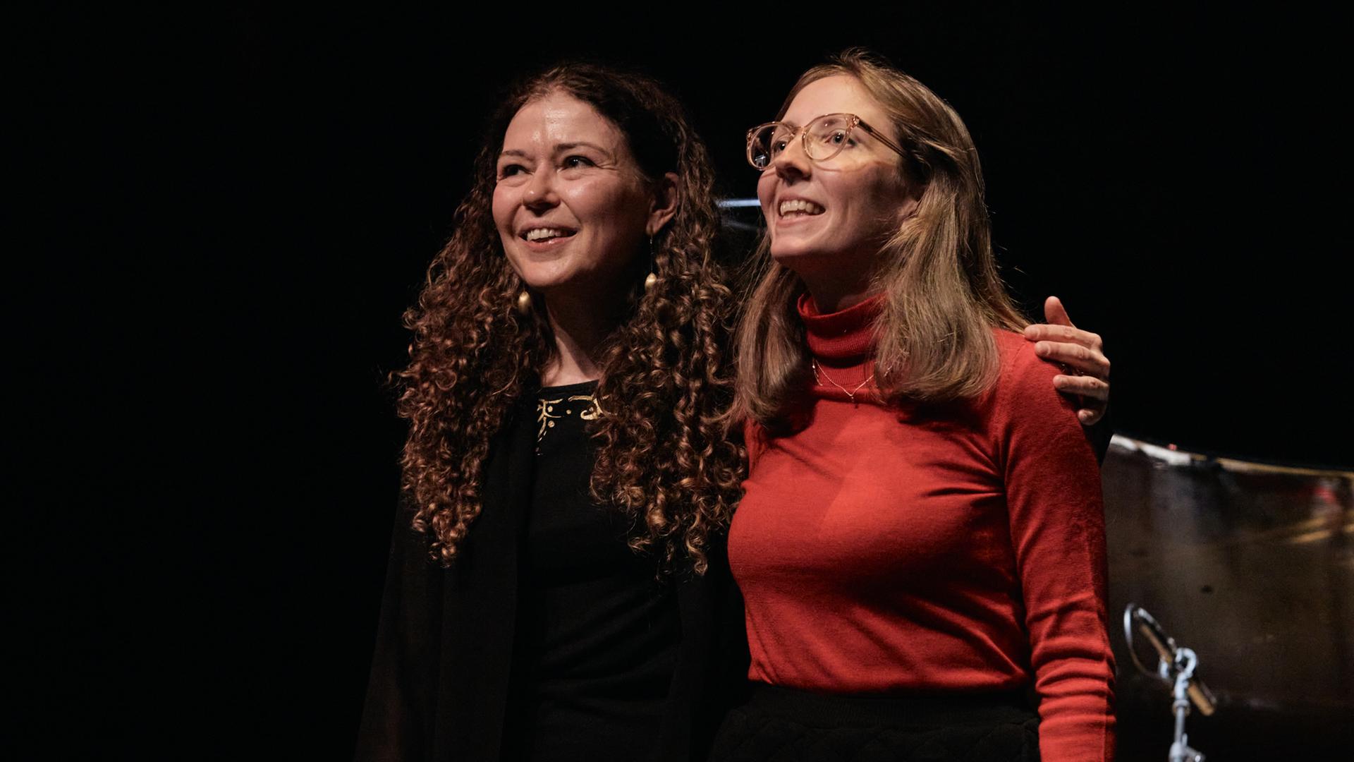 Zwei Frauen stehen eng nebeneinander auf einer dunklen Bühne und nehmen den Beifall des Publikums lächelnd entgegen.