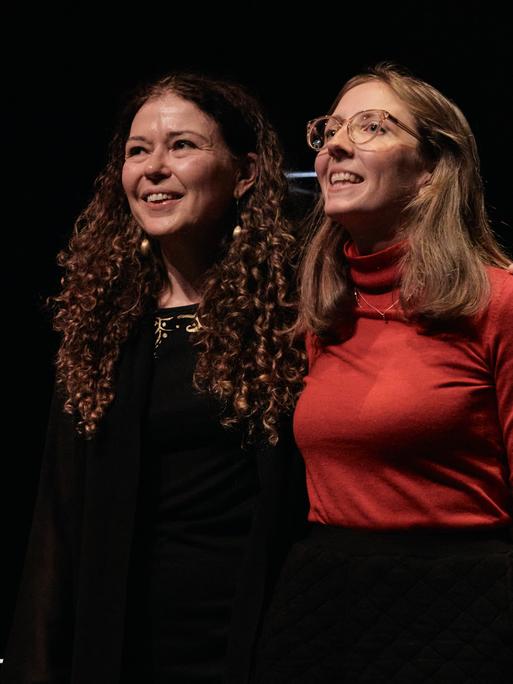 Zwei Frauen stehen eng nebeneinander auf einer dunklen Bühne und nehmen den Beifall des Publikums lächelnd entgegen.