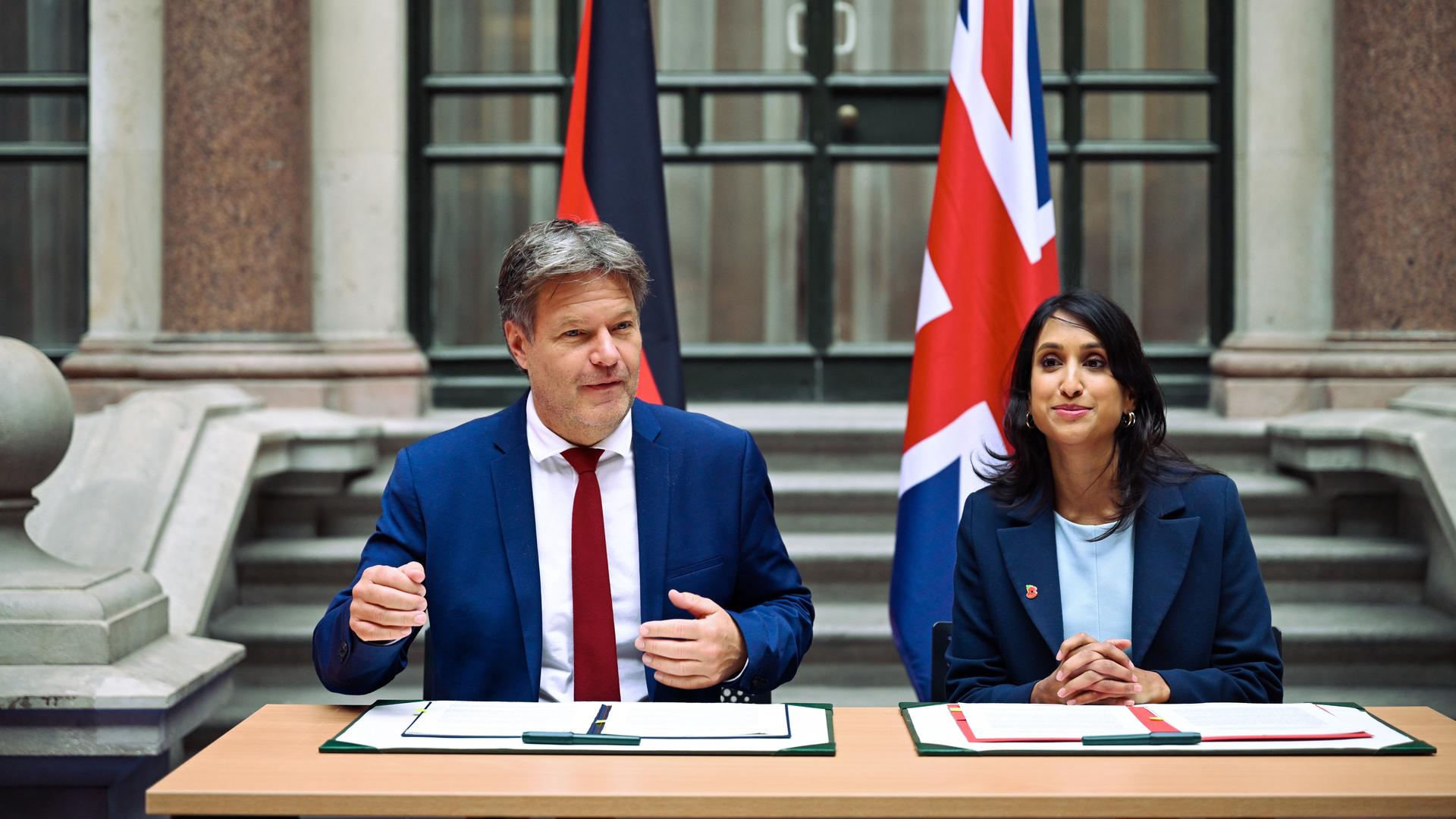 Eine Frau und ein Mann sitzen vor einem Tisch, auf dem Unterlagen liegen. Im Hintergrund hängen eine britische und eine deutsche Fahne.