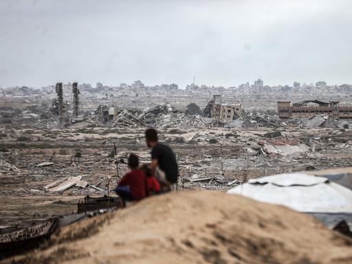 Zwei Personen, die von hinten zu sehen sind, darunter ein Kind, sitzen auf einem Hügel und schauen auf die zerstörte Stadt von Gaza