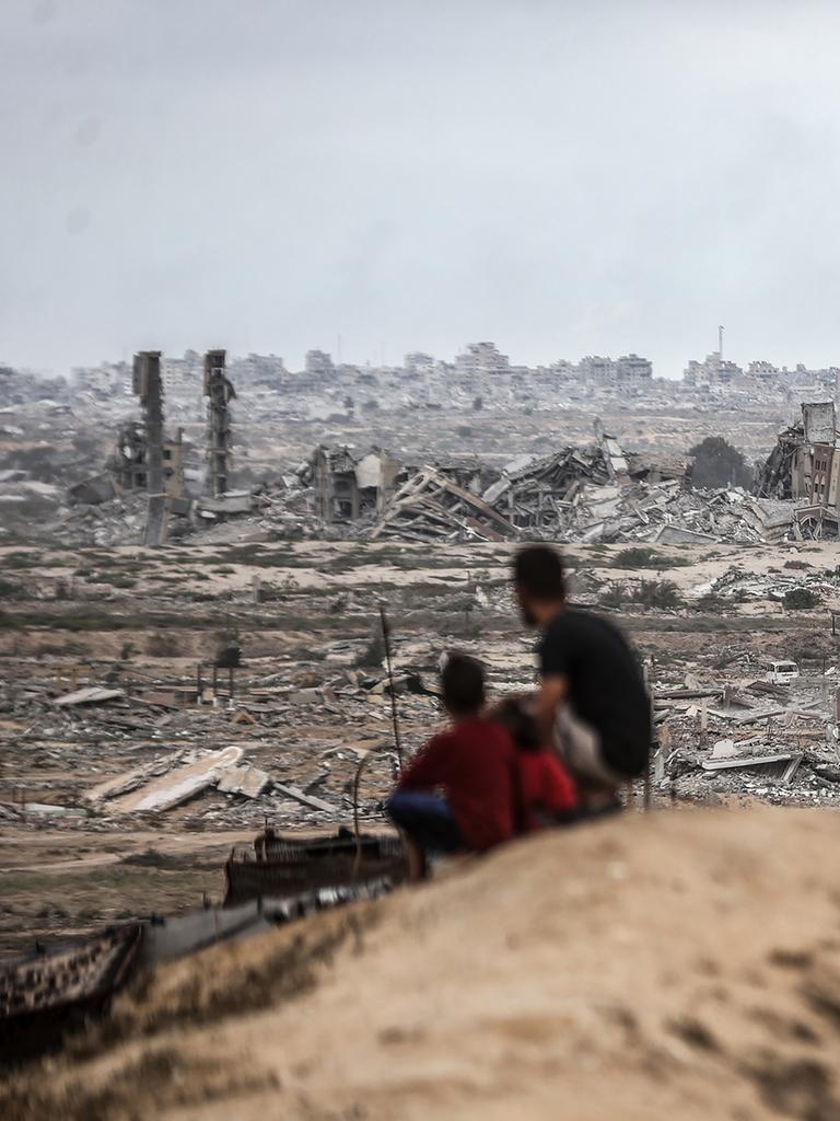 Zwei Personen, die von hinten zu sehen sind, darunter ein Kind, sitzen auf einem Hügel und schauen auf die zerstörte Stadt von Gaza
