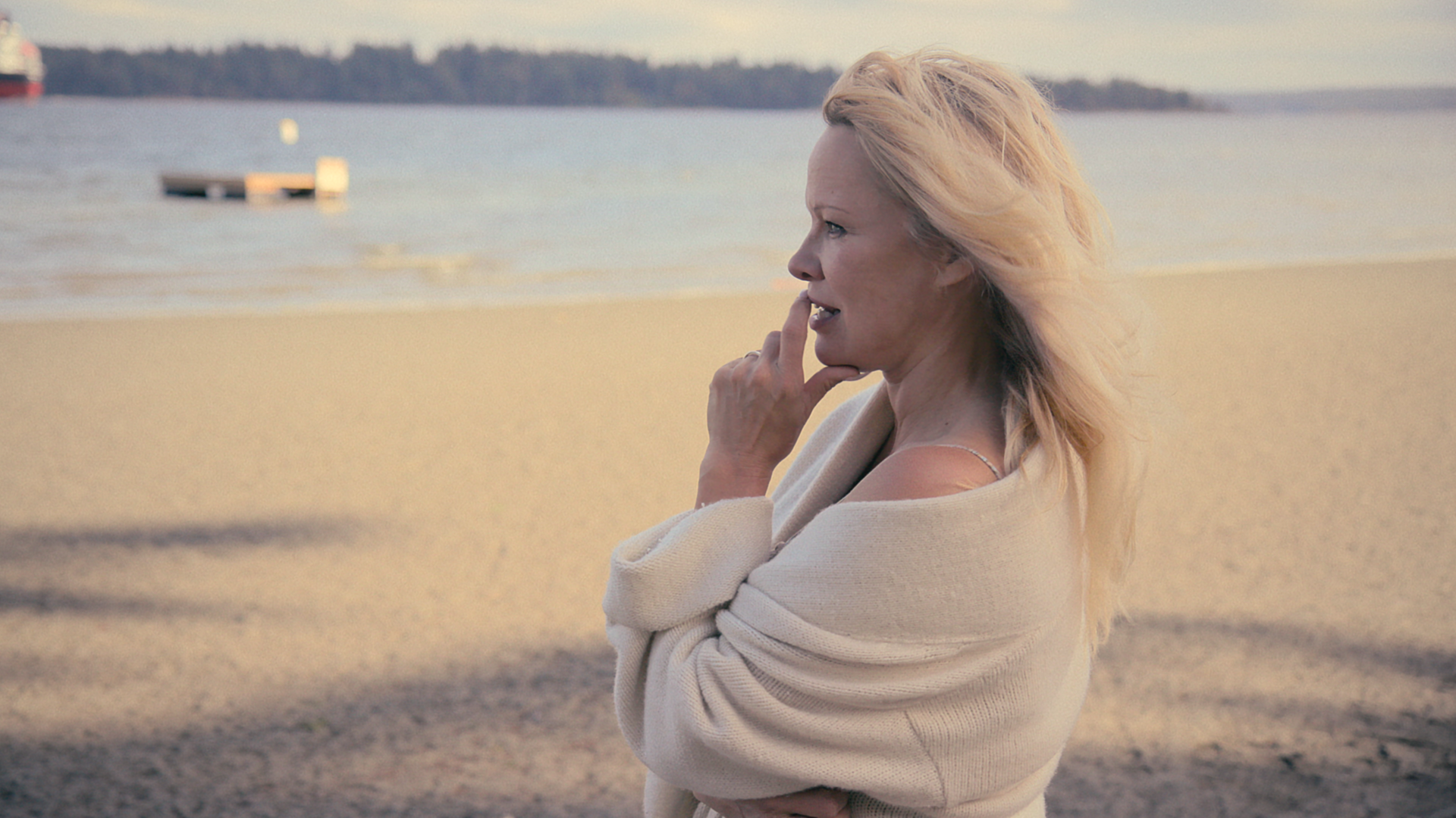 Die Schauspielerin Pamela Anderson steht an einem Sandstrand und schaut aufs Wasser. 