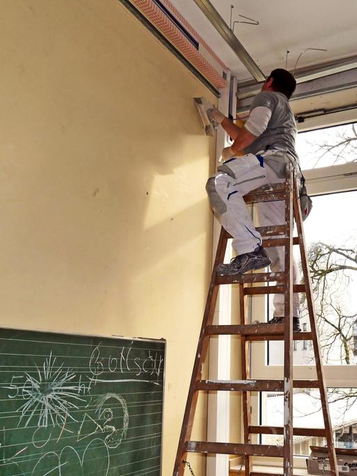 In einem Klassenzimmer steht ein Maler auf der Leiter und streicht die Wand.