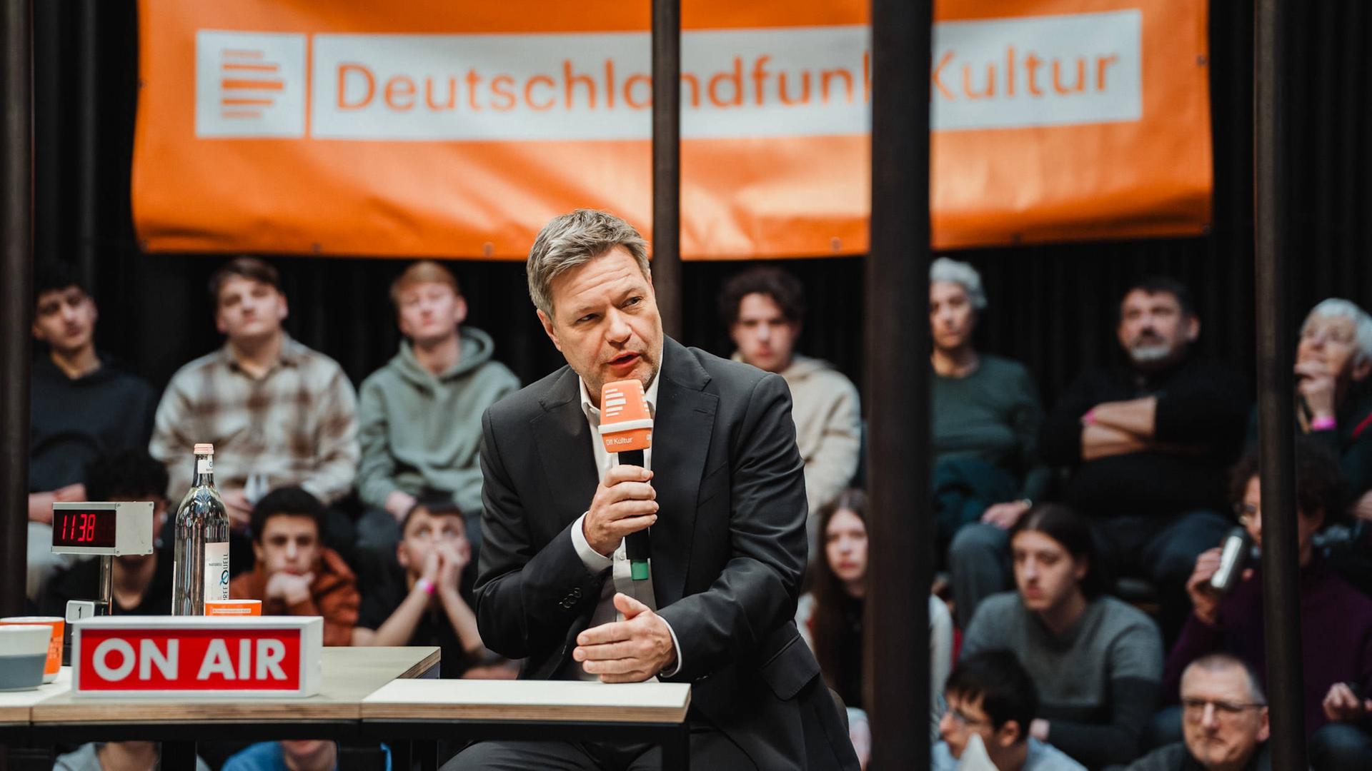 Bundeswirtschaftsminister Robert Habeck (Grüne) sitzt auf einem Podium und spricht ins Mikrofon. Im Hintergrund ein Live-Publikum und ein Banner mit dem Logo von Deutschlandfunk Kultur.