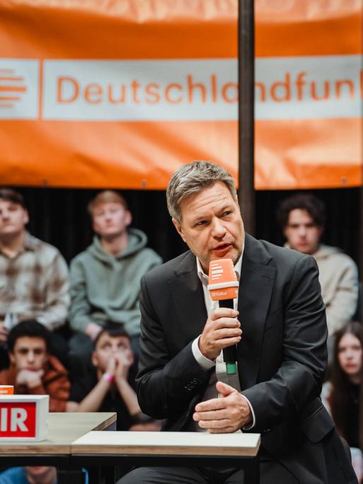 Bundeswirtschaftsminister Robert Habeck (Grüne) sitzt auf einem Podium und spricht ins Mikrofon. Im Hintergrund ein Live-Publikum und ein Banner mit dem Logo von Deutschlandfunk Kultur.