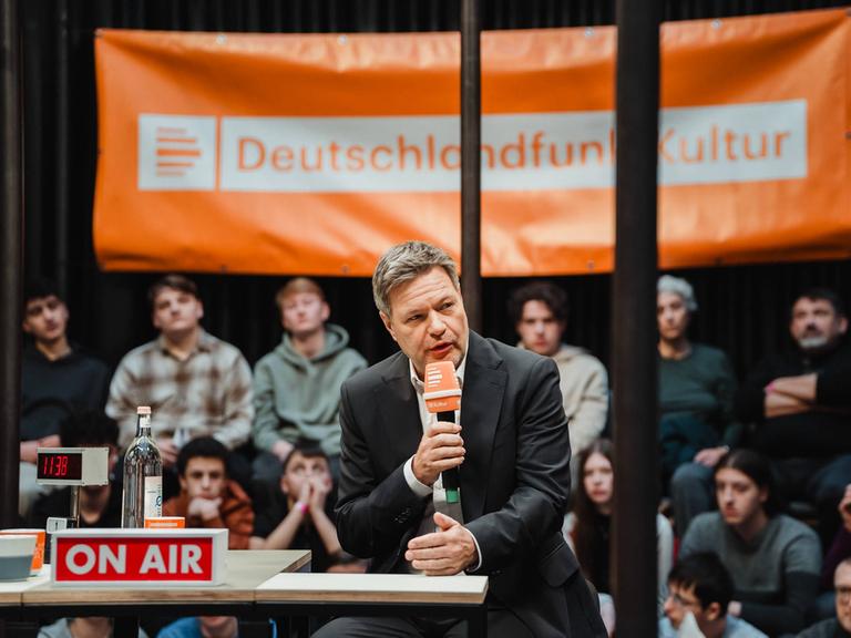 Bundeswirtschaftsminister Robert Habeck (Grüne) sitzt auf einem Podium und spricht ins Mikrofon. Im Hintergrund ein Live-Publikum und ein Banner mit dem Logo von Deutschlandfunk Kultur.