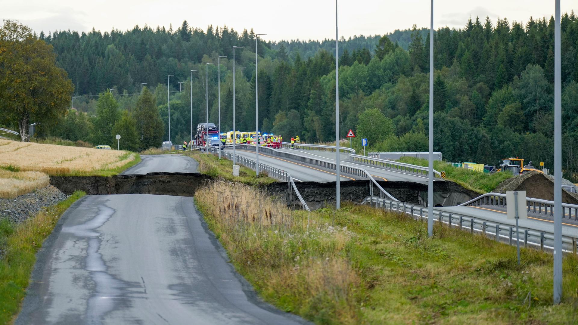 Auf dem Foto ist eine beschädigte Straße zu sehen. Auch die daneben liegende Eisenbahnstrecke ist durch den Erdrutsch unbenutzbar geworden. Im Hintergrund stehen Einsatzfahrzeuge von Feuerwehr und Polizei.