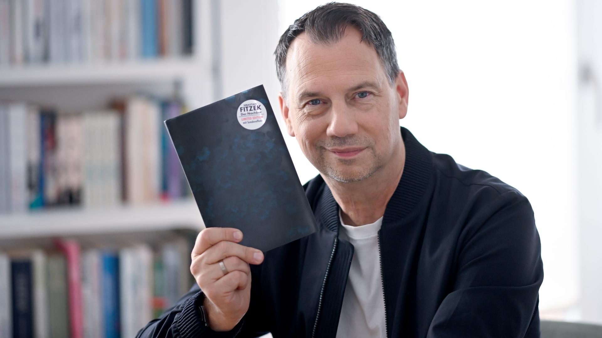 Sebastian Fitzek hält ein Buchcover hoch, auf dessen Rückseite ein Button mit seinem Namen klebt. Sebastian Fitzek hält ein Buchcover hoch, auf dessen Rückseite ein Button mit seinem Namen klebt.