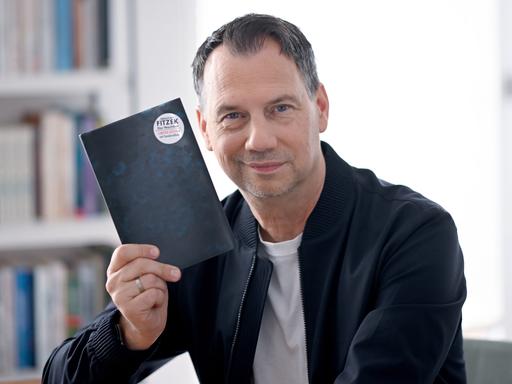 Sebastian Fitzek hält ein Buchcover hoch, auf dessen Rückseite ein Button mit seinem Namen klebt.