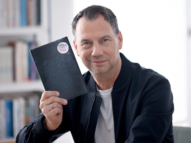 Sebastian Fitzek hält ein Buchcover hoch, auf dessen Rückseite ein Button mit seinem Namen klebt.