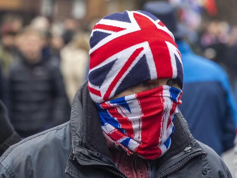 Ein Mann vermummt sich mit der englischen Union Jack, Mütze und Mundschutz. Demonstranten versammeln sich am 25. Januar 2026 in Crowborough, auf einem ehemaligen Militärgelände, in dem Asylanten untergebracht werden sollen.