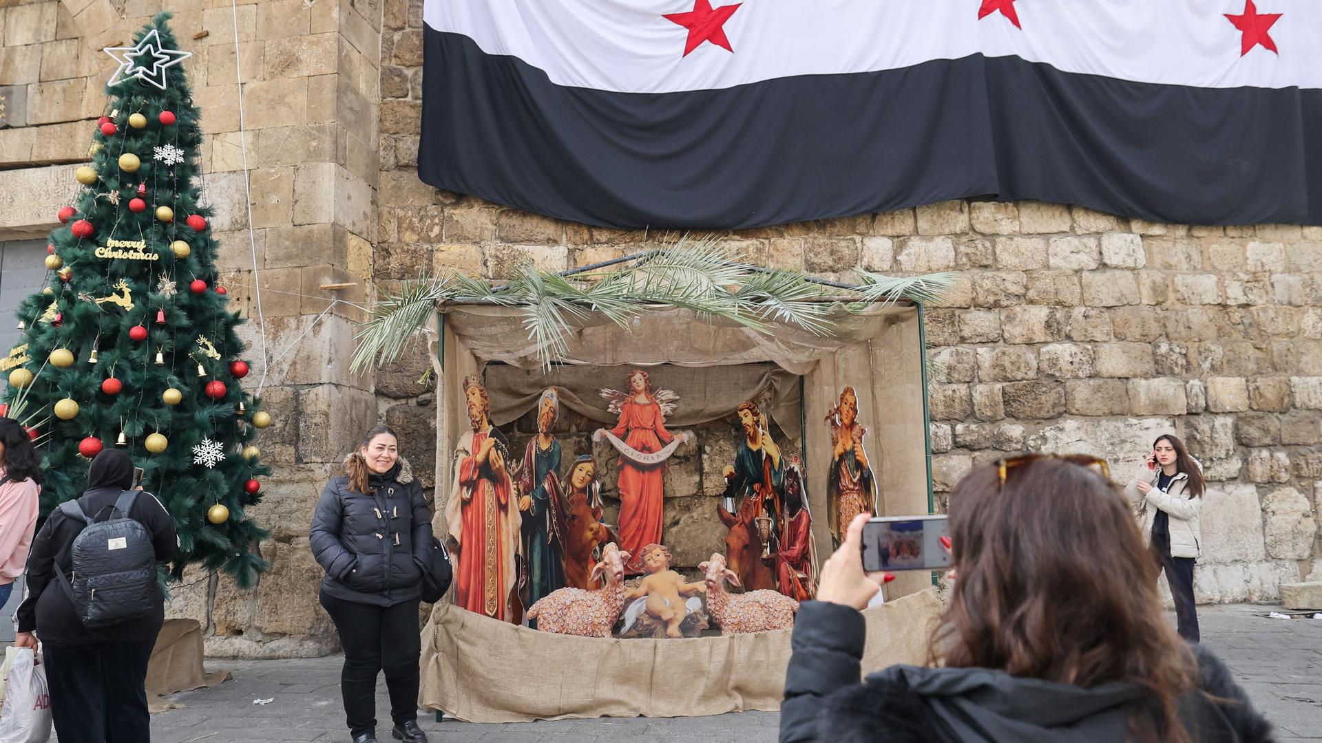 Eine Frau posiert vor einer weihnachtlichen Krippe, an einer Wand dahinter hängt eine syrische Flagge.