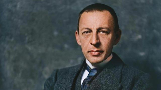 Sergej Rachmaninow sitzt vor einem grauen Hintergrund in aufgerichteter Pose auf einem Stuhl und blickt ernst in die Kamera.