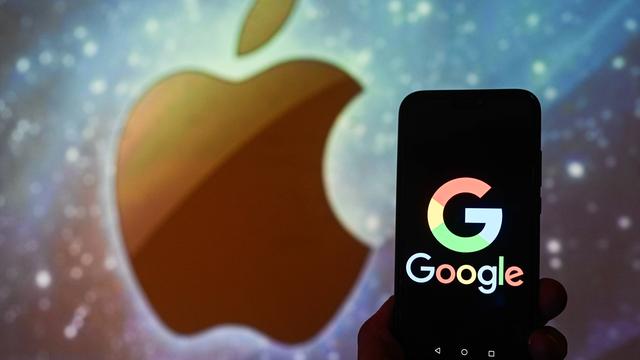 Das Logo des US-Technologieunternehmens Google wird auf einem Smartphone angezeigt, im Hintergrund abgebildet ist das Apfel-Logo des amerikanischen Konzerns Apple.