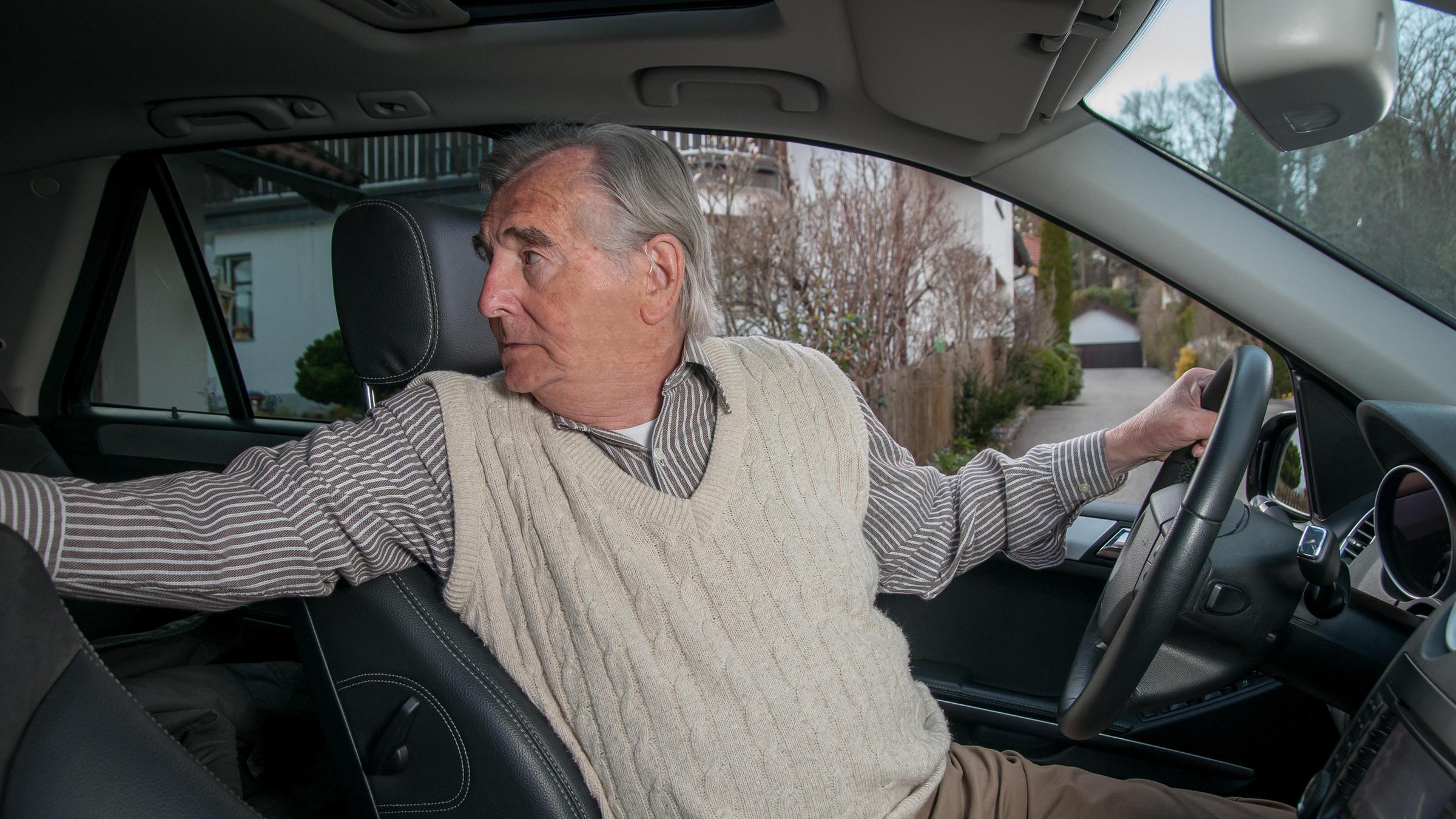 Ein älterer Mann sitzt am Steuer seines Wagens und dreht sich nach hinten, um rückwärts fahren zu können.