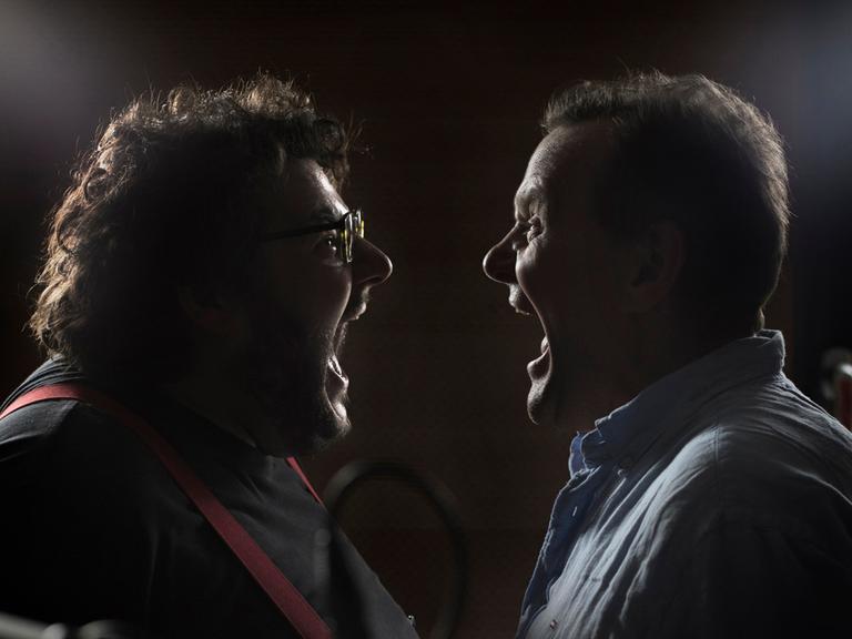 Devid Striesow und Axel Ranisch stehen sich in einem dunklen Raum im Profil gegenüber und schreien sich mit weit aufgerissenen Mündern an.