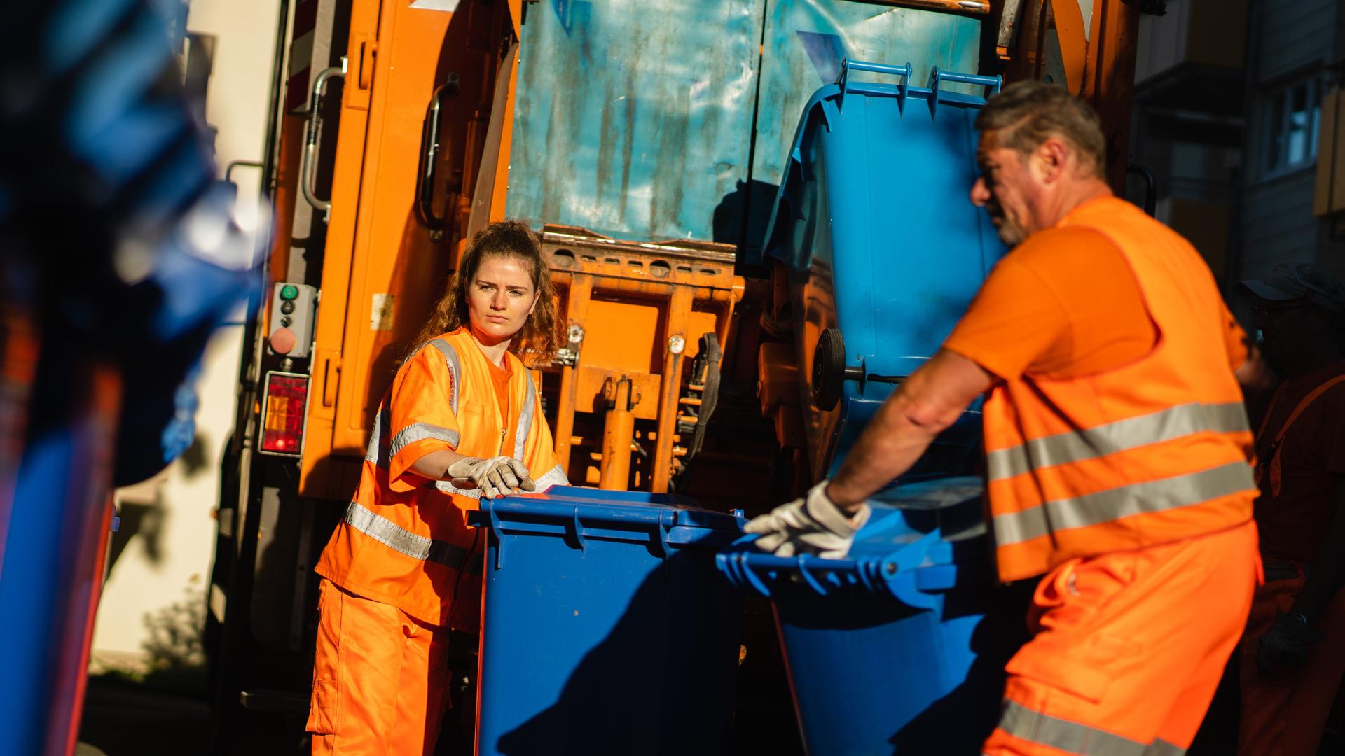 Zwei Müllwerker bei der Arbeit: Eine junge Frau und ein mittelalter Mann schieben Papiermülltonnen zum Laster der Müllabfuhr
