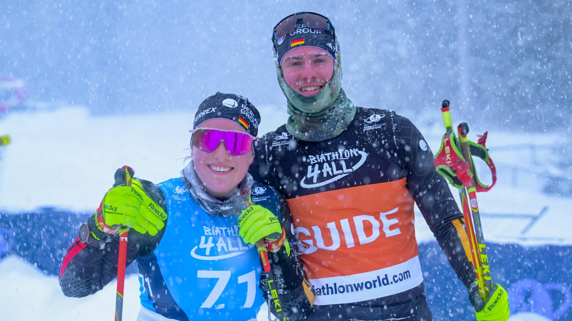 Eine Sportlerin und ein Sportler im Schneegestöber, sie steht links und trägt eine pinke, verspiegelte Sportsonnenbrille. Beide haben Skistöcke in den Händen und lächeln in die Kamera. 