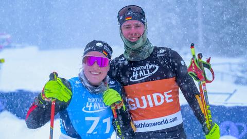 Eine Sportlerin und ein Sportler im Schneegestöber, sie steht links und trägt eine pinke, verspiegelte Sportsonnenbrille. Beide haben Skistöcke in den Händen und lächeln in die Kamera. 