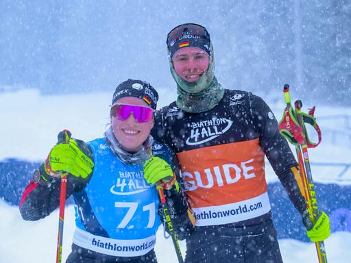 Eine Sportlerin und ein Sportler im Schneegestöber, sie steht links und trägt eine pinke, verspiegelte Sportsonnenbrille. Beide haben Skistöcke in den Händen und lächeln in die Kamera. 