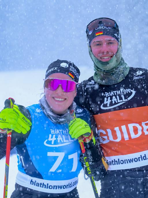 Eine Sportlerin und ein Sportler im Schneegestöber, sie steht links und trägt eine pinke, verspiegelte Sportsonnenbrille. Beide haben Skistöcke in den Händen und lächeln in die Kamera. 
