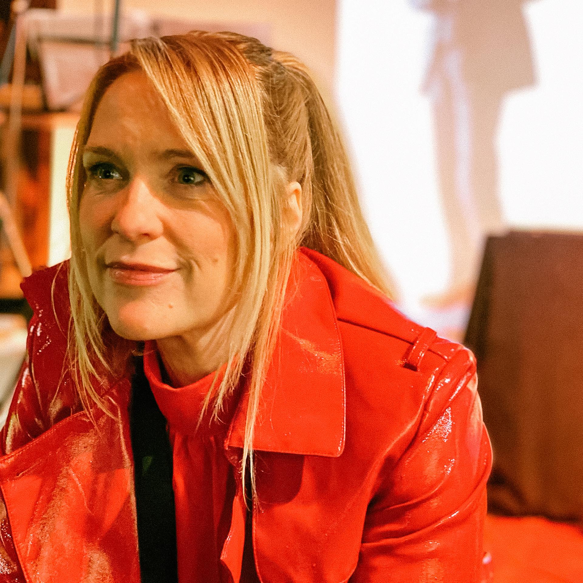 Porträt von Mayjia Gille live beim Rilke Programm. Sie trägt einen roten Mantel aus Lackleder und ihr glattes, blondes Haar in einem Pferdeschwanz.