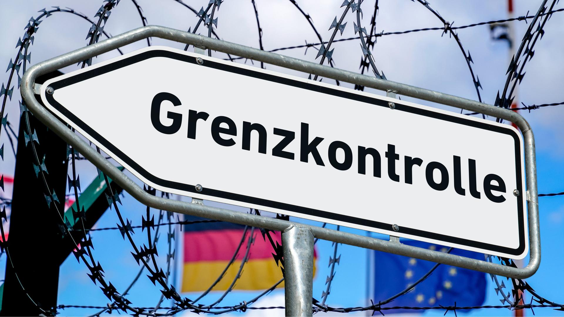 Ein weißer Wegweiser mit der Aufschrift "Grenzkontrolle" vor einem Zaun, gesichert mit Stacheldraht und den wehenden Flaggen der Bundesrepublik Deutschland und der Europäischen Union im Hintergrund. (Montage)