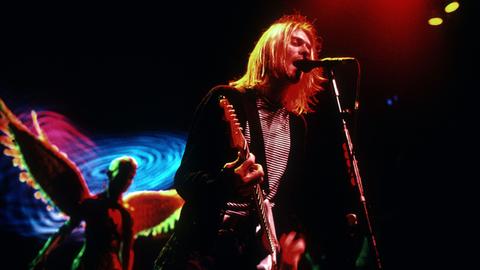 Kurt Cobain steht mit Gitarre auf einer schummrig ausgeleuchteten Bühne und singt.