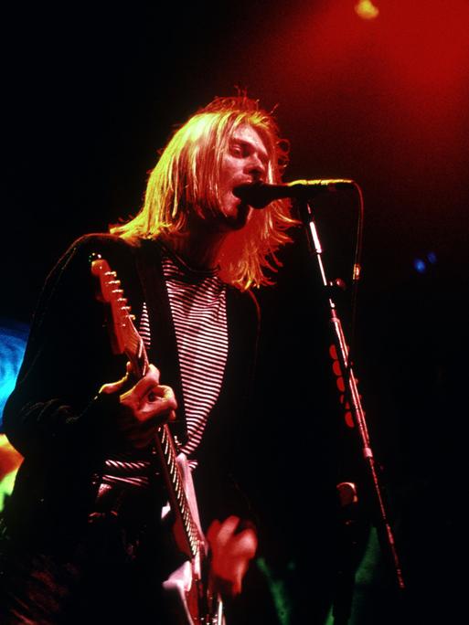 Kurt Cobain steht mit Gitarre auf einer schummrig ausgeleuchteten Bühne und singt.