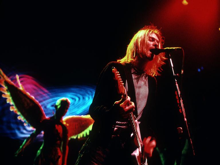 Kurt Cobain steht mit Gitarre auf einer schummrig ausgeleuchteten Bühne und singt.