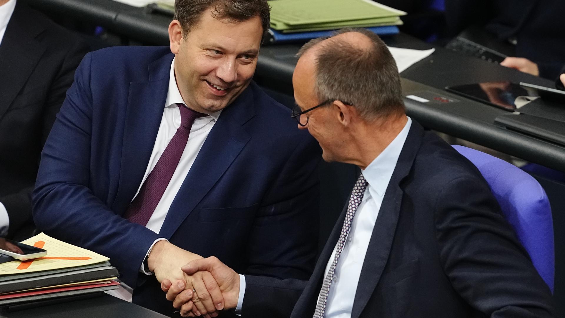SPD-Chef Lars Klingbeil und Bundeskanzler Friedrich Merz (CDU) schütteln sich lächelnd die Hand. Sie sitzen im Bundestag.