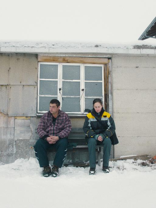 Marco und Anna sitzen auf einer Bank vor einem Haus. Um sie herum ist die Landschaft weiß und verschneit. Beide schauen eher missmutig drein. 