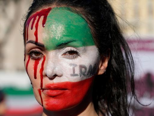 Nahaufnahme einer Frau bei einer Protestkundgebung. Sie hat die eine Hälfte ihres Gesichtes mit den Farben der iranischen Flagge bemalt, die andere Hälfte mit Blutstropfen.