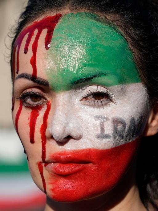 Nahaufnahme einer Frau bei einer Protestkundgebung. Sie hat die eine Hälfte ihres Gesichtes mit den Farben der iranischen Flagge bemalt, die andere Hälfte mit Blutstropfen.