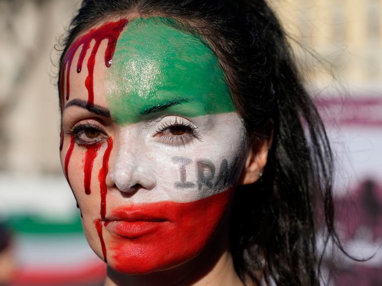 Nahaufnahme einer Frau bei einer Protestkundgebung. Sie hat die eine Hälfte ihres Gesichtes mit den Farben der iranischen Flagge bemalt, die andere Hälfte mit Blutstropfen.
