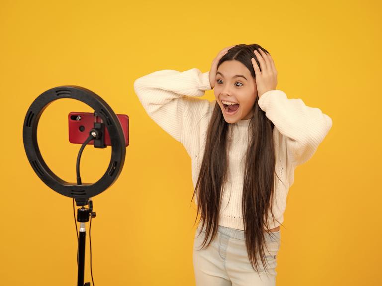 Eine junge Bloggerin vor dem Kameraphone mit weit aufgerissenen Augen vor gelbem Hintergrund.