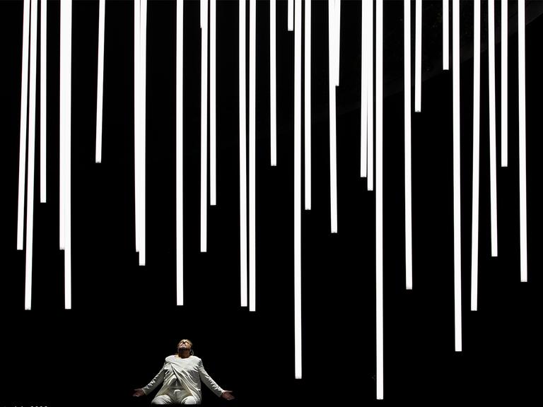 Ein Mann im weißen Anzug kniet auf einer völlig schwarzen Bühne, das Gesicht nach oben gewendet, von dort fallen weiße Lichtsäulen auf ihn herab.