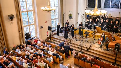 Ein großer Saal  mit hoher Decke und hohen Fenstern. Von der Decke hängen große Leuchter. Das Parkett ist mit roten Stühlen ausgestattet. Auf der Bühne stehen schwarz gekleidete Sängerinnen und Sänger und Musikerinnen und Musiker.