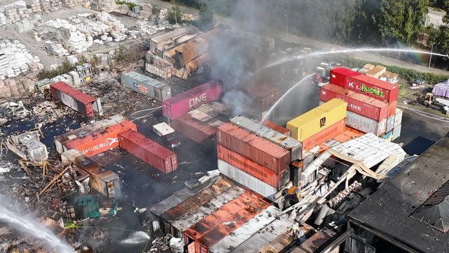 Blick aus der Luft auf einen Teil von dem Hafen in Hamburg: Qualmende Trümmer und Erde neben bunten Containern. Von der Seite löscht die Feuer-Wehr mit Wasser.