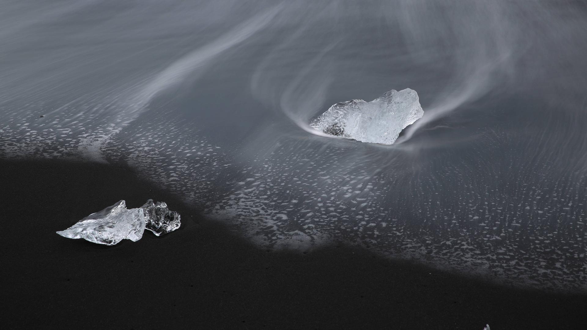 Zwei Eisblöcke umweht von eisigem Wind und Schnee auf einem schwarzen Grund. Sie liegen in schwarzem Wasser.