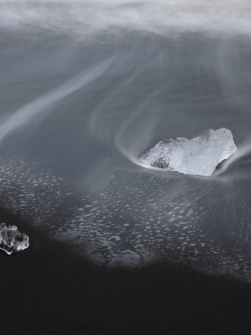 Zwei Eisblöcke umweht von eisigem Wind und Schnee auf einem schwarzen Grund. Sie liegen in schwarzem Wasser.