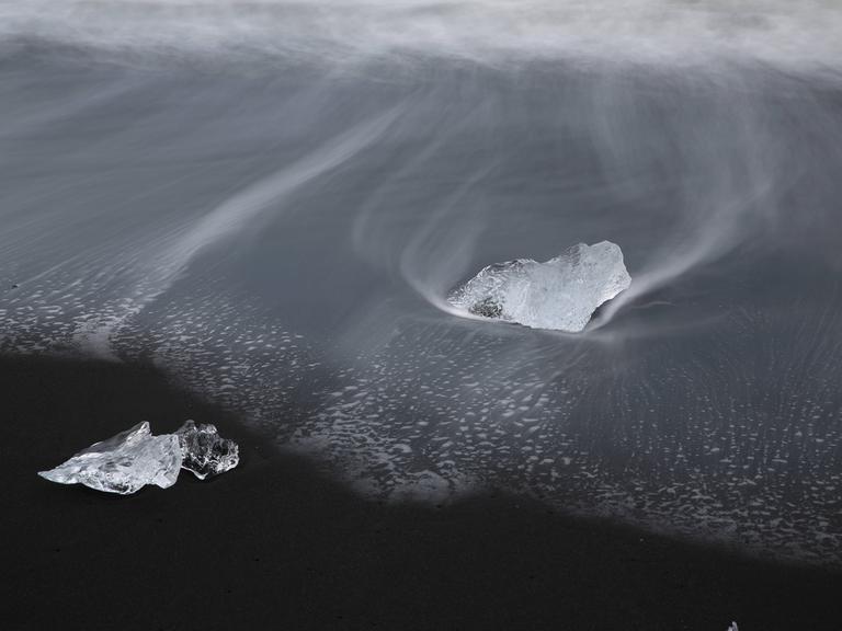 Zwei Eisblöcke umweht von eisigem Wind und Schnee auf einem schwarzen Grund. Sie liegen in schwarzem Wasser.