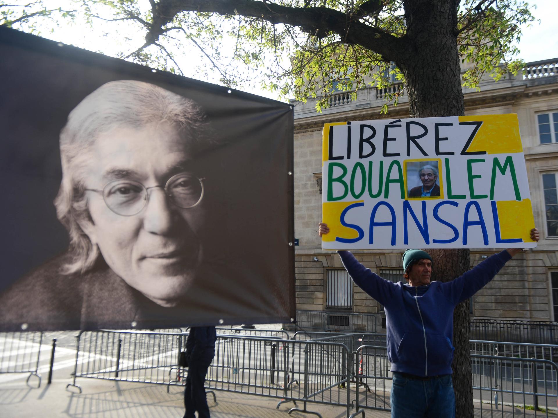 Auf der Straße protestiert ein Mann mit einem Banner für die Freilassung von Boualem Sansal, links daneben ein Plakat, das den franko-algerischen Schriftsteller zeigt.